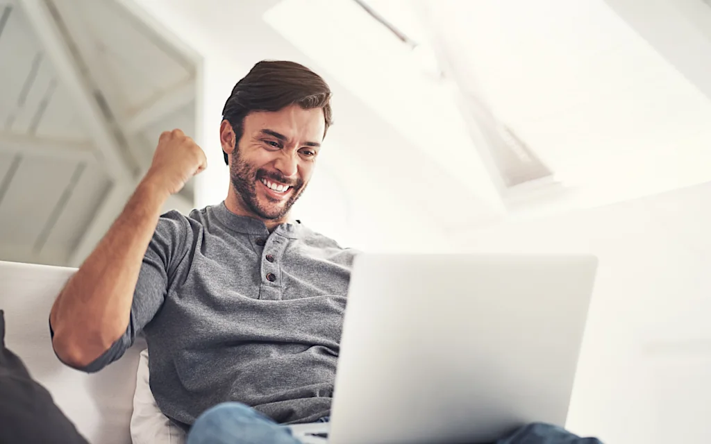 Mann mit lächelndem Gesicht sitzt am Laptop und ballt vor Freude seine Faust