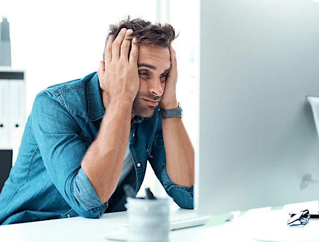 Ein verzweifelter und überforderter Mann im blauen Hemd sitzt vor dem Computer, starrt auf den Bildschir und hält den Kopf in seinen Händen.